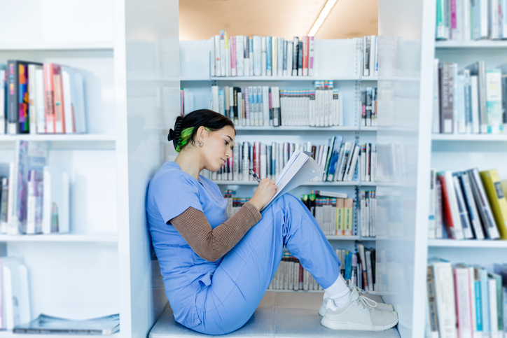 A nurse wearing scrubs marks up a document while sitting in a library.