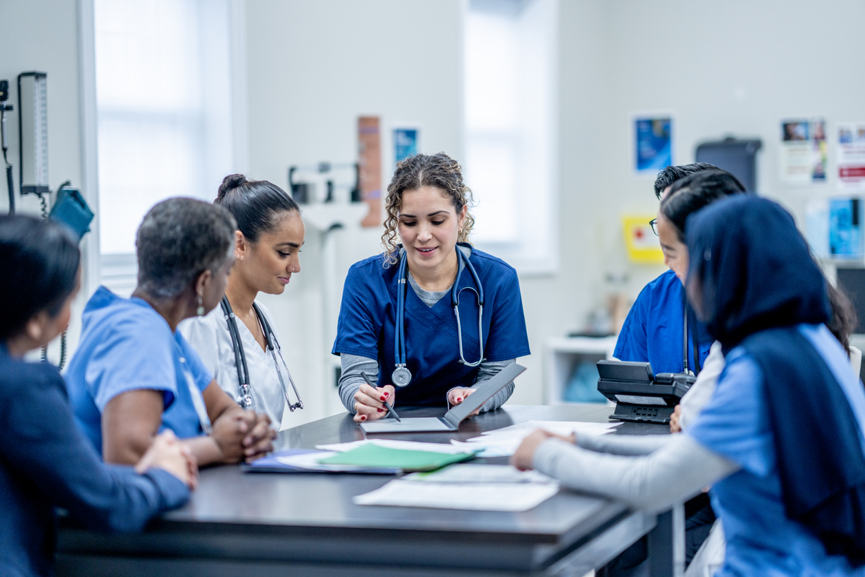 A nurse shows a tablet to a group of colleagues seated around a table.