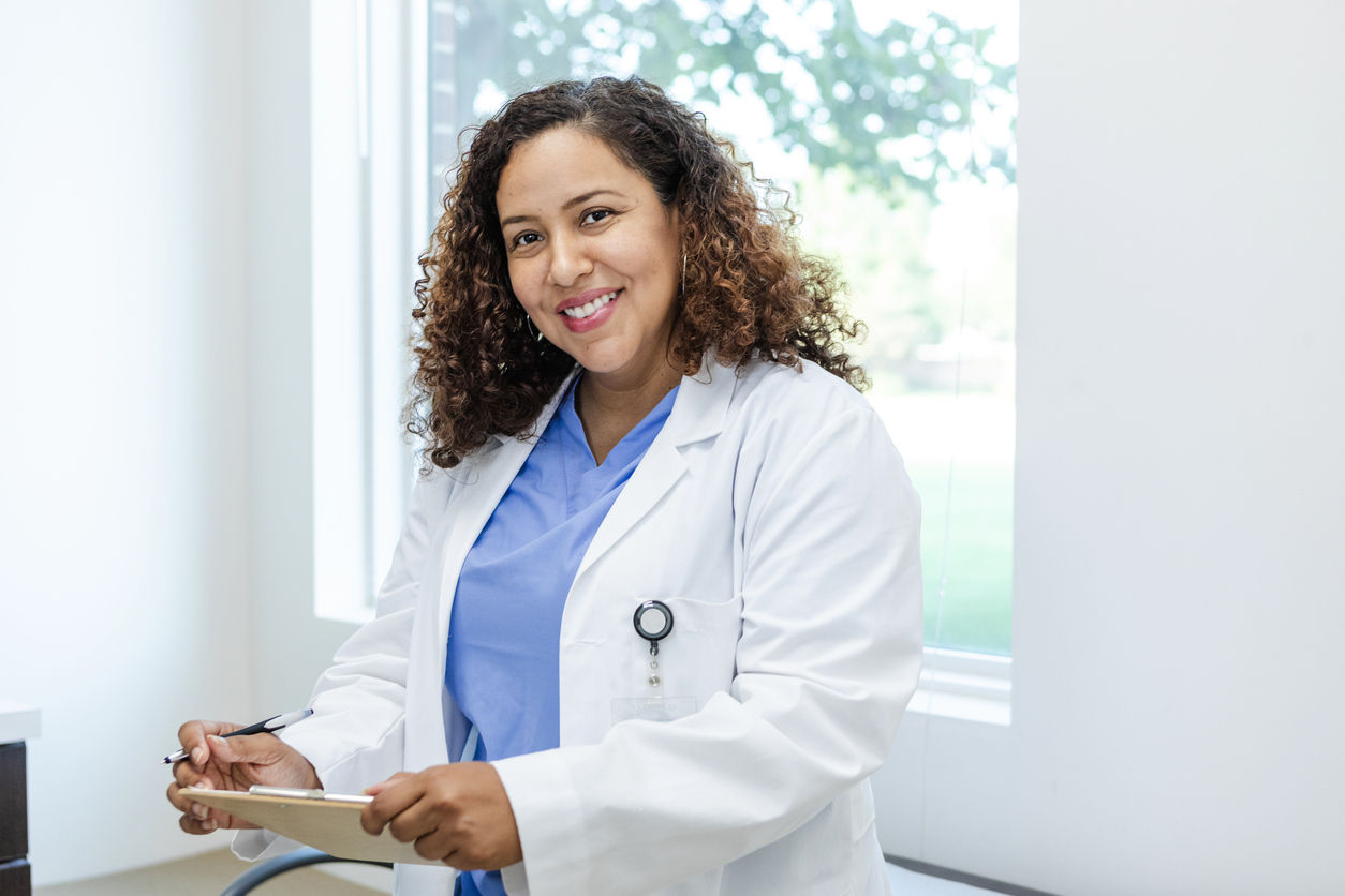 A nurse leader making notes on a clipboard pauses to smile at the camera.