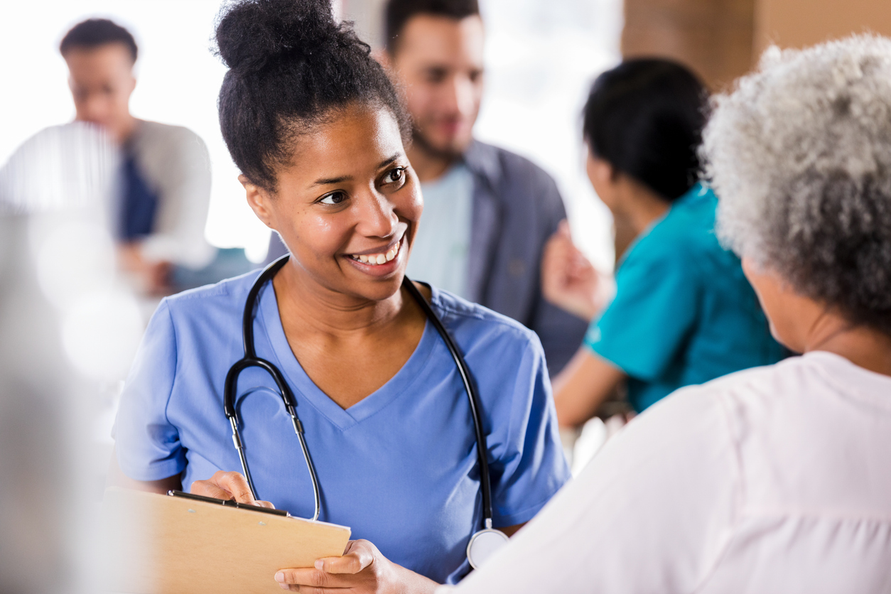 A nurse wearing scrubs and holding a clipboard speaks to a patient.