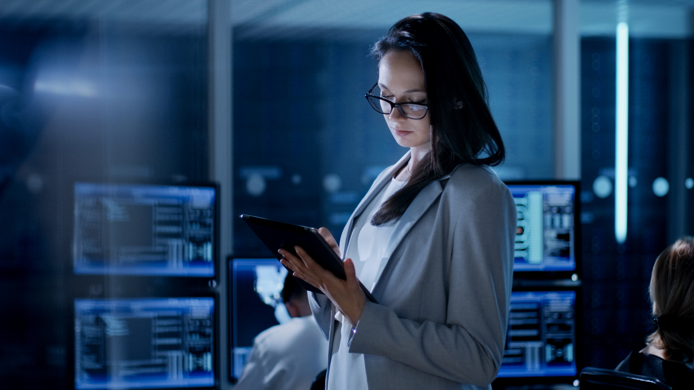 A Cybersecurity Professional Stands Near Several Computer Monitors Looking at a Tablet.