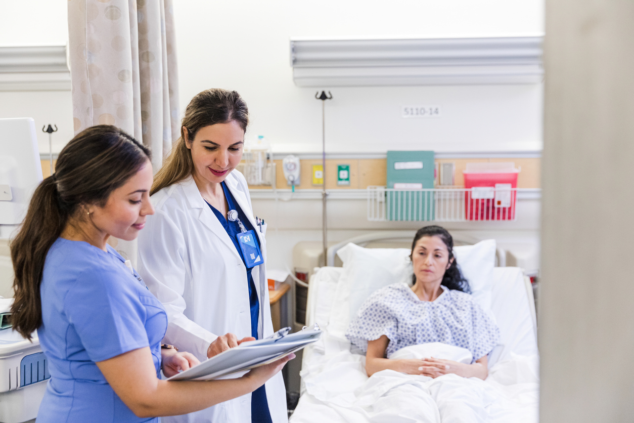 A Student Nurse Takes Notes Regarding a Patient in a Hospital Bed While a Nurse Practitioner Observes..jpg